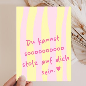 Könnte beinhalten: Eine Grußkarte mit gelb-rosa gestreiftem Hintergrund. Die Karte zeigt den Text "Du kannst soooooooo stolz auf dich sein." in Pink, mit einem rosa Herz am unteren Rand. Die Karte wird von einer Hand gehalten.
