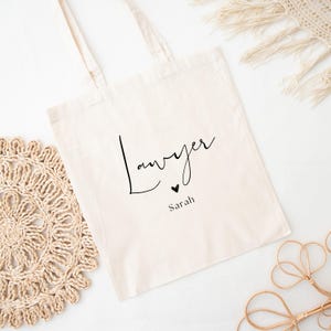 May include: Cream-colored tote bag with the word "Lawyer" in a black script font, a small black heart, and the name "Sarah" below. The bag has two handles and is made of a canvas-like material. The background includes decorative elements.