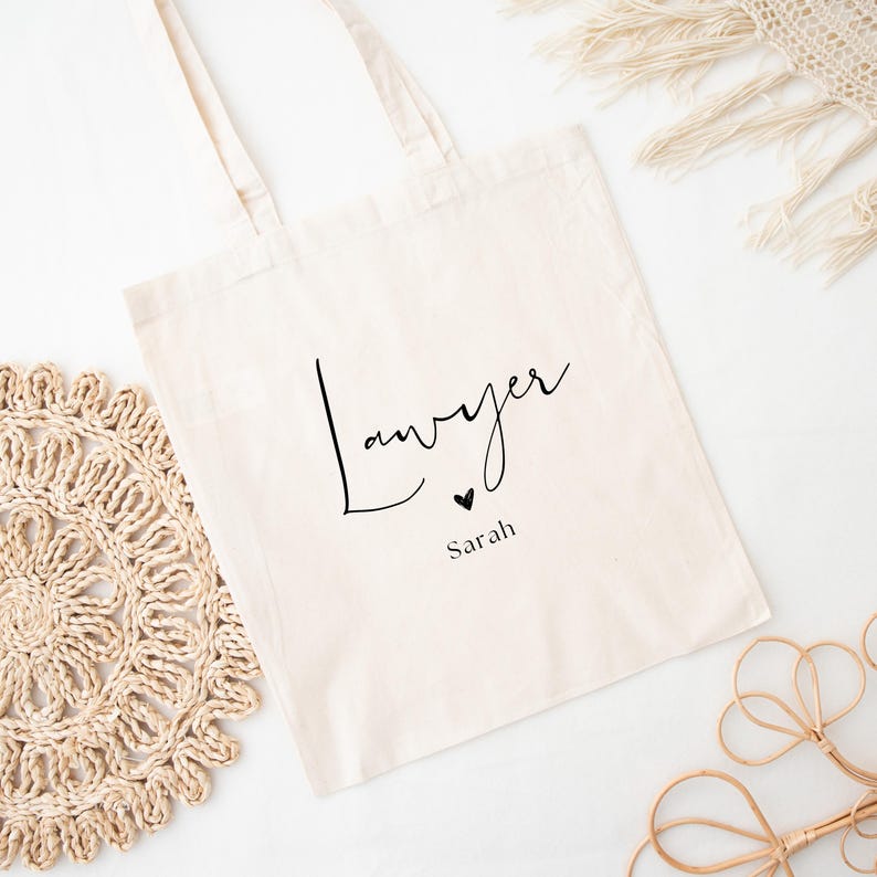 May include: Cream-colored tote bag with the word "Lawyer" in a black script font, a small black heart, and the name "Sarah" below. The bag has two handles and is made of a canvas-like material. The background includes decorative elements.