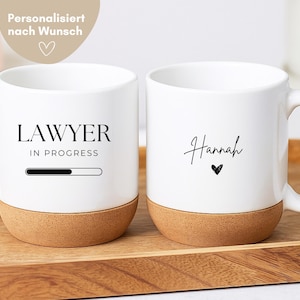 Könnte beinhalten: Zwei weiße Keramiktassen mit Korkboden. Eine Tasse zeigt den schwarzen Schriftzug "LAWYER IN PROGRESS" über einer Ladebalkengrafik. Die andere Tasse trägt den Namen "Hannah" in schwarzer Schreibschrift über einem kleinen schwarzen Herz.