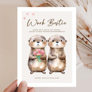 Puede incluir: Tarjeta de felicitación con la frase "Work Bestie" y una ilustración en acuarela de dos nutrias. Una nutria sostiene un ramo de flores rosas. La tarjeta también incluye texto en alemán. El fondo es beige neutro.