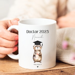 May include: White ceramic mug featuring a black and brown otter wearing a graduation cap and holding a diploma. The mug reads "Doctor 2025" and "You Did It" with the name "Hannah" written in cursive.