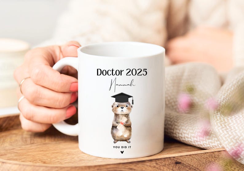 May include: White ceramic mug featuring a black and brown otter wearing a graduation cap and holding a diploma. The mug reads "Doctor 2025" and "You Did It" with the name "Hannah" written in cursive.