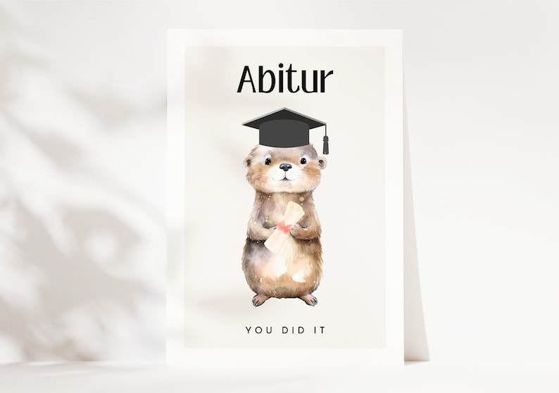 May include: A graduation card featuring a cartoon otter wearing a black graduation cap and holding a diploma. The card has the text "Abitur" and "You Did It" on it.