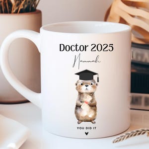 May include: White ceramic mug with a black handle. The mug features a black and white illustration of a cartoon otter wearing a graduation cap and holding a diploma. The text "Doctor 2025" and "Hannah" are printed in black on the mug. The text "You Did It" is printed in black at the bottom of the mug.