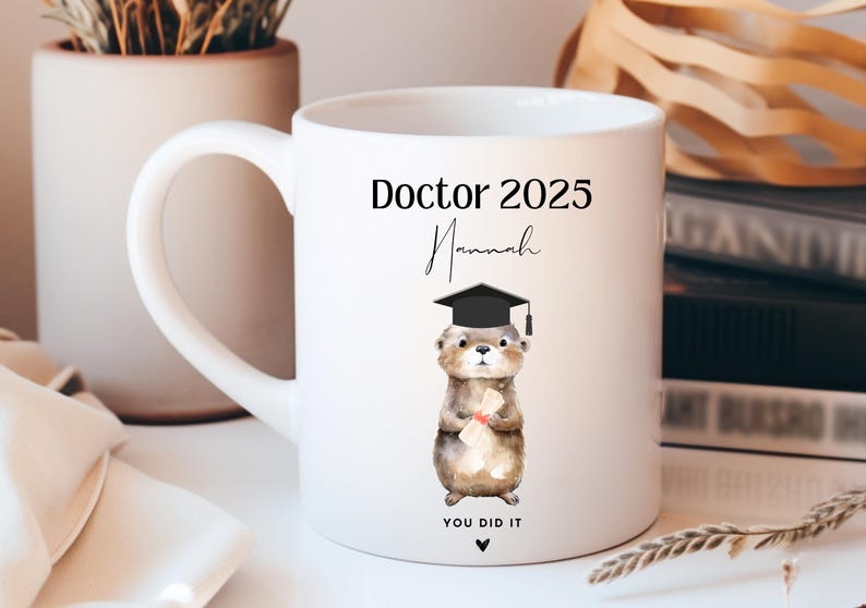 May include: White ceramic mug with a black handle. The mug features a black and white illustration of a cartoon otter wearing a graduation cap and holding a diploma. The text "Doctor 2025" and "Hannah" are printed in black on the mug. The text "You Did It" is printed in black at the bottom of the mug.