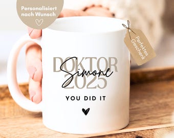 May include: White ceramic mug with a black and brown design that says "Doktor Simone 2023 You Did It". The mug has a pink handle and a small heart at the bottom.