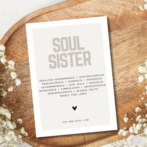 May include: A beige card with the words "Soul Sister" in large, bold, gray letters. The card has a list of words describing a soul sister, such as "absolute heart person", "home protector", "problem solver", "listener", "advisor", "crisis manager", "good soul", "counselor", "cheerleader", "confidant", "supporter", "tear dryer", "better half", and "person for life". The card also has a small black heart and the words "I love you" in black text.