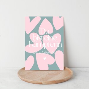 Pu&ograve; includere: Una stampa artistica rettangolare con uno sfondo verde acqua e forme di cuore rosa. La stampa presenta un testo bianco che recita "Du bist eine K&auml;mpferin, du schaffst es." La stampa &egrave; esposta su un vassoio di legno.