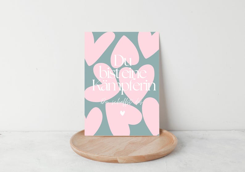 Pu&ograve; includere: Una stampa artistica rettangolare con uno sfondo verde acqua e forme di cuore rosa. La stampa presenta un testo bianco che recita "Du bist eine K&auml;mpferin, du schaffst es." La stampa &egrave; esposta su un vassoio di legno.