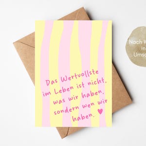 May include: A greeting card with a yellow and pink striped design. The card has the German text "Das Wertvollste im Leben ist nicht, was wir haben, sondern wen wir haben." A brown envelope is behind the card.