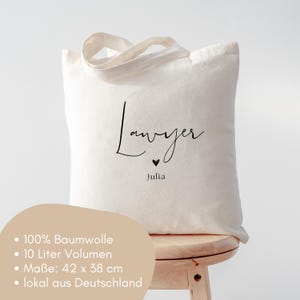 May include: Cream-colored tote bag with the word "Lawyer" in a handwritten font, a small heart, and the name "Julia" below. The bag has a simple design with a handle and is made of a soft material.
