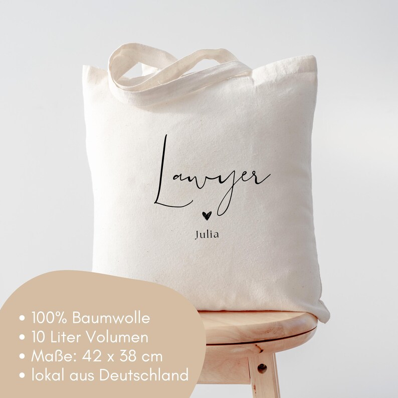 May include: Cream-colored tote bag with the word "Lawyer" in a handwritten font, a small heart, and the name "Julia" below. The bag has a simple design with a handle and is made of a soft material.
