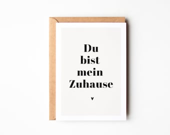 Op de afbeelding: Een witte kaart met een bruine envelop. De kaart heeft zwarte tekst die "Du bist mein Zuhause" luidt met een klein hartje onderaan.