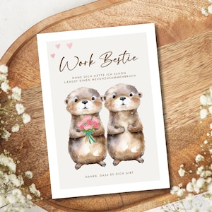 May include: A greeting card with the text "Work Bestie" and two otter illustrations. One otter holds a bouquet of pink flowers. The card has a light beige background with small pink hearts and text in German.