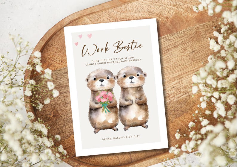 May include: A greeting card with the text "Work Bestie" and two otter illustrations. One otter holds a bouquet of pink flowers. The card has a light beige background with small pink hearts and text in German.