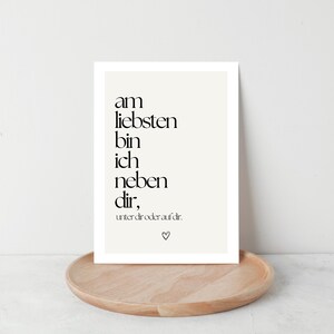 May include: A framed print with black text on a cream background, resting on a wooden tray. The text reads, "am liebsten bin ich neben dir," with a small heart symbol. The print is displayed against a white wall.