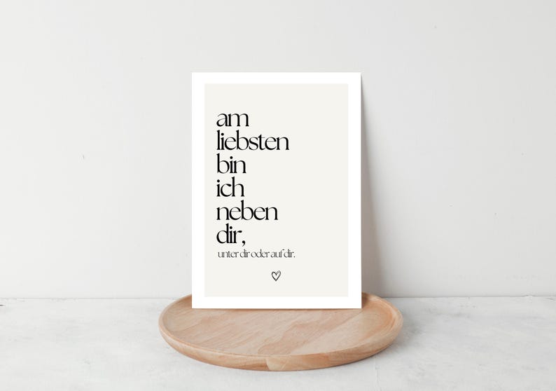 May include: A framed print with black text on a cream background, resting on a wooden tray. The text reads, "am liebsten bin ich neben dir," with a small heart symbol. The print is displayed against a white wall.