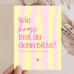 Könnte beinhalten: Eine rechteckige Grußkarte mit einem vertikalen, wellenförmigen Streifenmuster in Hellgelb und Hellrosa. Der Text lautet "Wie krass bist du denn bitte?" und "Du kannst so stolz auf dich sein." in Pink, mit einem kleinen pinken Herzsymbol unten.