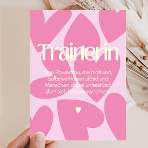 Puede incluir: Una tarjeta rectangular con fondo rosa claro y varias formas de corazón en rosa oscuro. El texto "Trainerin" aparece en letras grandes de color crema, seguido de un texto en alemán que describe a una entrenadora que motiva.
