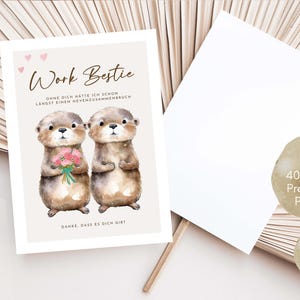 May include: A greeting card with the text "Work Bestie" and two otter illustrations. One otter holds a bouquet of pink flowers. The card is accompanied by a blank white card and two informational stickers.