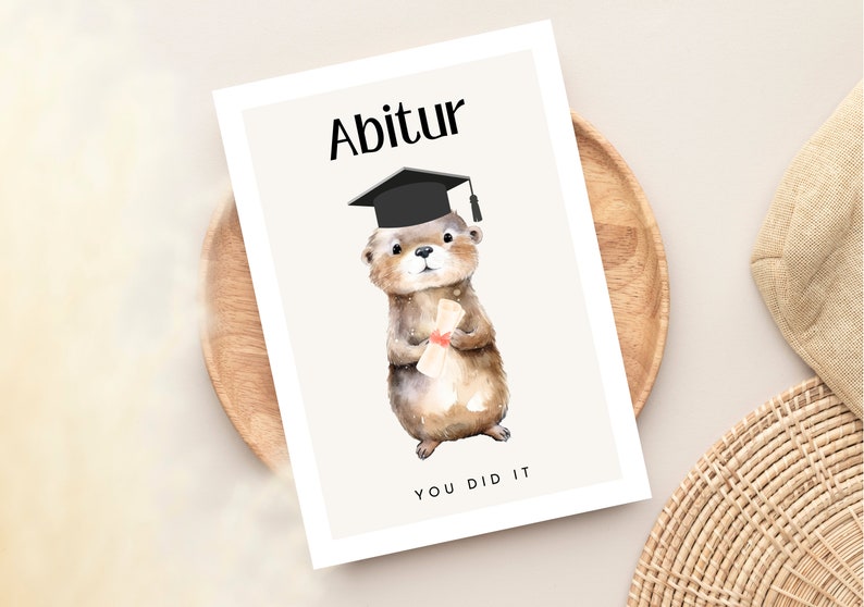 May include: A graduation card featuring a cute otter wearing a black graduation cap and holding a diploma. The card has the text "Abitur" and "You Did It" on it.