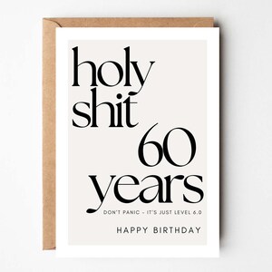 May include: A birthday card with the words "holy shit 60 years" in black font. The card also includes the phrases "Don't panic - it's just level 6.0" and "Happy Birthday". The card is in a white frame with a brown envelope.