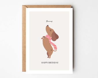 Puede incluir: Tarjeta de felicitación con un dibujo de un perro salchicha marrón con una bufanda rosa con corazones. El texto "Boooof" está encima del perro y "HAPPY BIRTHAY" debajo, sobre un fondo claro.