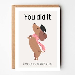 May include: A graduation greeting card featuring a dachshund wearing a graduation cap and a pink scarf with hearts. The card reads "You did it" at the top and "HERZLICHEN GL&Uuml;CKWUNSCH" at the bottom. The card is in a brown envelope.