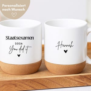 May include: Two white ceramic mugs with cork bases. The left mug features the black text "Staatsexamen 2026 You did it" with a small heart. The right mug features the name "Hannah" in script font with a small heart below it.