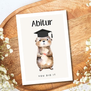 May include: A graduation card featuring a cute otter wearing a black graduation cap and holding a diploma. The card has the text "Abitur" and "You Did It" on it.
