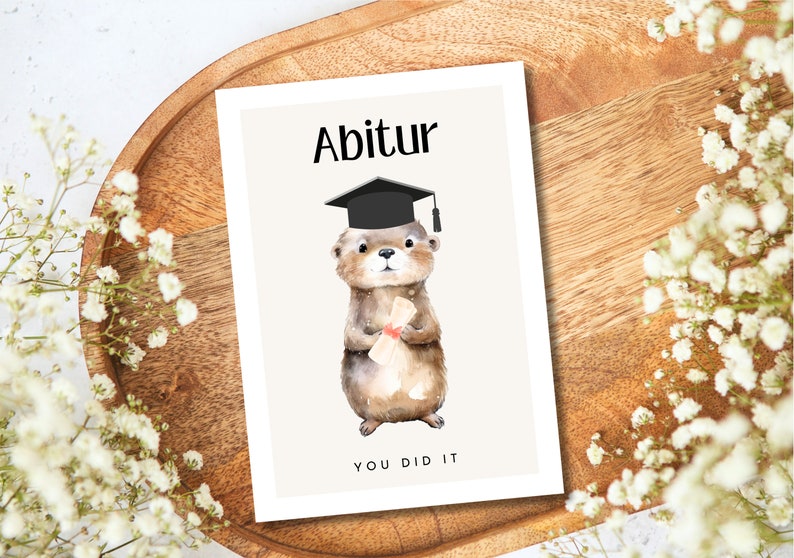 May include: A graduation card featuring a cute otter wearing a black graduation cap and holding a diploma. The card has the text "Abitur" and "You Did It" on it.