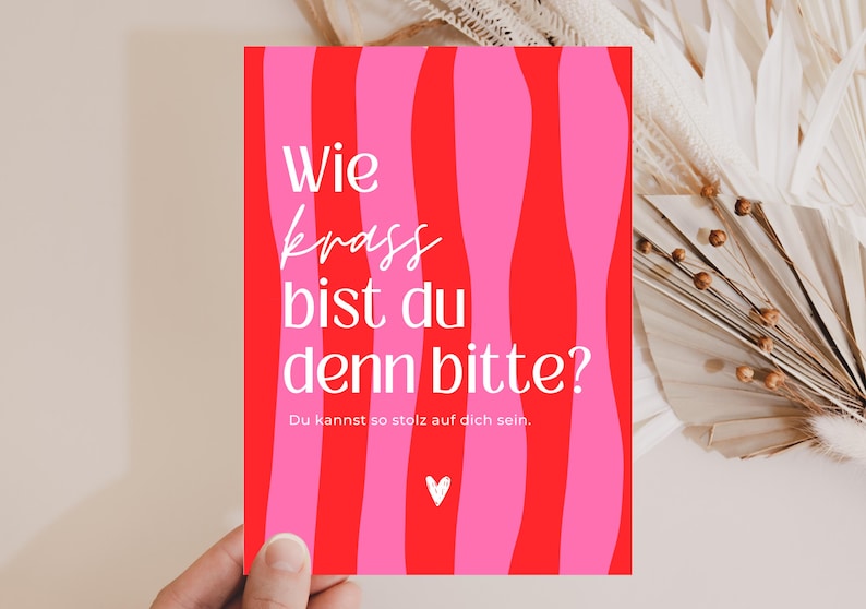 May include: A rectangular greeting card featuring a vertical wavy striped pattern in alternating shades of bright pink and red. The card displays the white text "Wie krass bist du denn bitte?" followed by "Du kannst so stolz auf dich sein." and a small white heart icon.