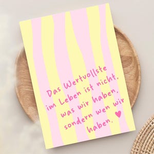 May include: A greeting card with a yellow and pink striped pattern. The card features the German text "Das Wertvollste im Leben ist nicht, was wir haben, sondern wen wir haben" in pink, with a pink heart at the bottom.