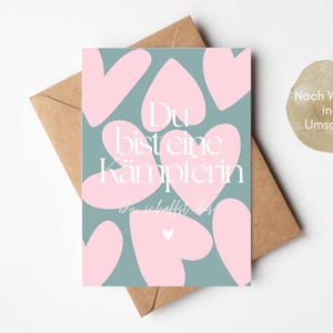 Pu&ograve; includere: Un biglietto di auguri con sfondo verde acqua e forme di cuore rosa. Il biglietto riporta il testo tedesco "Du bist eine K&auml;mpferin, Du schaffst das" in bianco. Il biglietto &egrave; all'interno di una busta marrone.
