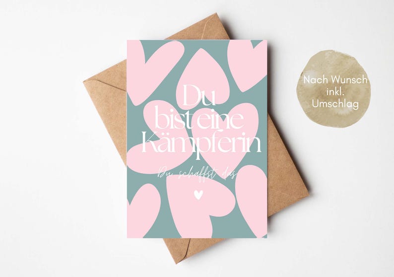 Pu&ograve; includere: Un biglietto di auguri con sfondo verde acqua e forme di cuore rosa. Il biglietto riporta il testo tedesco "Du bist eine K&auml;mpferin, Du schaffst das" in bianco. Il biglietto &egrave; all'interno di una busta marrone.