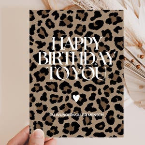 Könnte beinhalten: Eine rechteckige Geburtstagskarte mit einem braun-schwarzen Leopardenmuster. Der Text "HAPPY BIRTHDAY TO YOU" ist in großen weißen Großbuchstaben gedruckt, gefolgt von einem kleinen weißen Herz und dem Text "DU WUNDERVOLLER MENSCH".