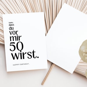 May include: A white card with black text that reads "Danke, dass du vor mir 50 wirst. Happy Birthday." The card is on a light brown background with a wooden fan behind it. The card is made of 400g/m2 premium paper and is a DIN A6 postcard.