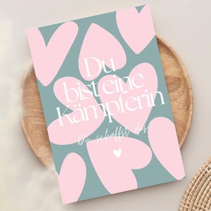 Pu&ograve; includere: Un biglietto di auguri con sfondo verde acqua e forme di cuore rosa. Il biglietto riporta il testo tedesco "Du bist eine K&auml;mpferin" in bianco, che si traduce in "Sei una combattente". Il biglietto &egrave; su un piatto di legno.