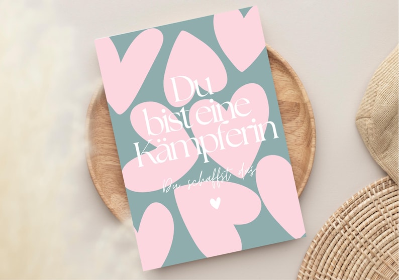 Pu&ograve; includere: Un biglietto di auguri con sfondo verde acqua e forme di cuore rosa. Il biglietto riporta il testo tedesco "Du bist eine K&auml;mpferin" in bianco, che si traduce in "Sei una combattente". Il biglietto &egrave; su un piatto di legno.