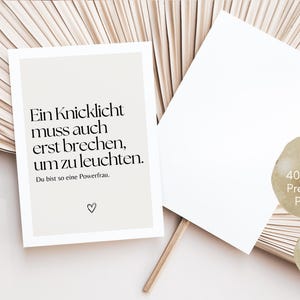 Puede incluir: Una postal blanca con una cita en blanco y negro en alem&aacute;n: "Ein Knicklicht muss auch erst brechen, um zu leuchten." La cita va seguida del texto "Du bist so eine Powerfrau." y un peque&ntilde;o s&iacute;mbolo de coraz&oacute;n. La postal est&aacute; sobre un fondo marr&oacute;n claro.