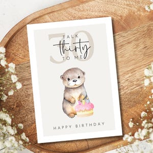 May include: A birthday card with a watercolor illustration of an otter holding a cake. The card reads "Talk Thirty to Me" and "Happy Birthday". The card is on a wooden tray with white flowers.