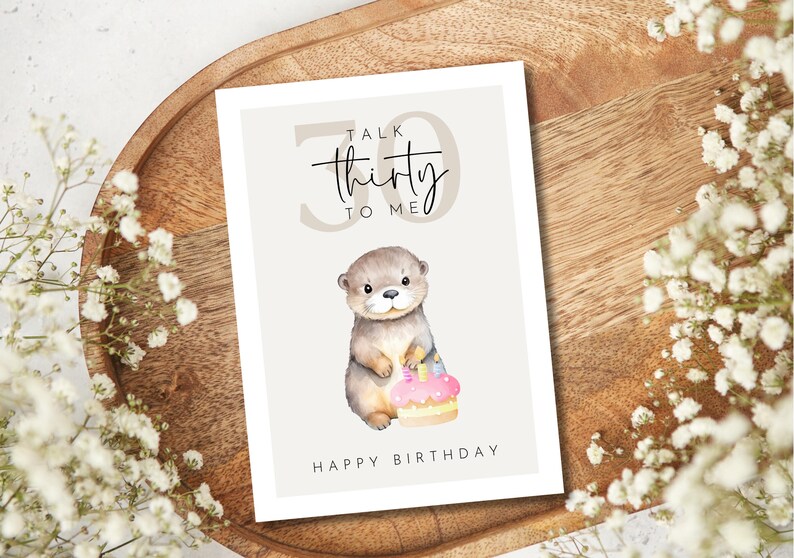 May include: A birthday card with a watercolor illustration of an otter holding a cake. The card reads "Talk Thirty to Me" and "Happy Birthday". The card is on a wooden tray with white flowers.