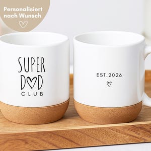 Pode incluir: Duas canecas de cerâmica branca com base de cortiça. A caneca da esquerda apresenta o texto preto SUPER DAD CLUB com um coração substituindo a letra A. A caneca da direita exibe o texto EST. 2026 acima de um pequeno coração. Ambas possuem alça.
