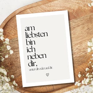 May include: A white card with black text that reads "am liebsten bin ich neben dir, unter dir oder auf dir." A small heart is at the bottom. The card is on a wooden tray, surrounded by white flowers.