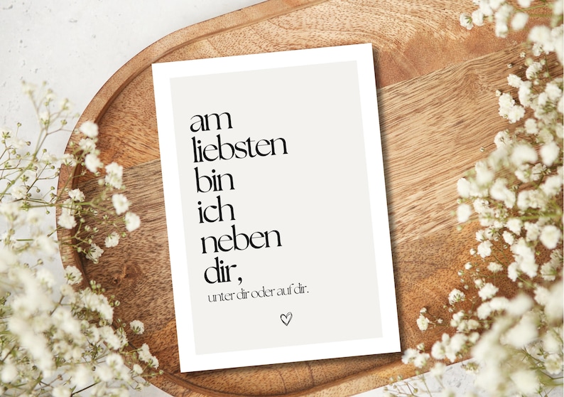 May include: A white card with black text that reads "am liebsten bin ich neben dir, unter dir oder auf dir." A small heart is at the bottom. The card is on a wooden tray, surrounded by white flowers.