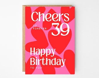 Könnte beinhalten: Geburtstagskarte mit rosa Hintergrund und roten Herzformen. Die Karte trägt die Aufschrift "Cheers to Forever 39" und "Happy Birthday to You" in weißem Text. Die Karte hat einen braunen Papierumschlag.