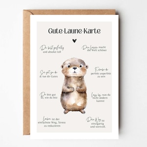 Puede incluir: Tarjeta de felicitación con una ilustración a la acuarela de una nutria. La tarjeta tiene texto en alemán, incluyendo "Gute-Laune-Karte" y frases inspiradoras. La nutria es marrón y gris, con una expresión linda.