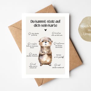 Puede incluir: Una tarjeta de felicitación con una ilustración de acuarela de una nutria. La tarjeta, con texto en alemán, está sobre un sobre marrón. También se incluye una pegatina con texto en alemán.