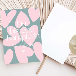 Pu&ograve; includere: Una cartolina con sfondo turchese e forme di cuore rosa. Il testo "Du bist eine K&auml;mpferin" &egrave; visualizzato in bianco. Accanto c'&egrave; una cartolina bianca vuota. La cartolina &egrave; realizzata in carta premium da 400 g/m2.